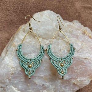 May include: A pair of teardrop-shaped earrings with a macrame design in a light green colour. The earrings are made with gold-coloured wire and have small gold beads.