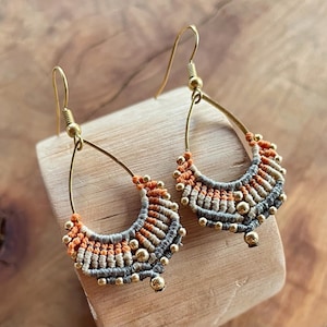 May include: A pair of teardrop-shaped earrings with a woven design in shades of grey, orange, and beige. The earrings are made with small gold beads and have a gold hook fastening.