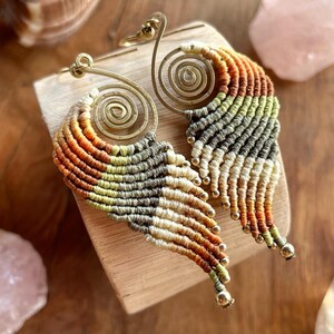 Peut inclure: Une paire de boucles d'oreilles faites à la main avec un motif en spirale et une frange de fil coloré. Les boucles d'oreilles sont réalisées dans une combinaison de tons dorés et bruns.