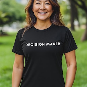 Puede incluir: Camiseta negra con el texto blanco "DECISION MAKER". La persona lleva la camiseta en un parque con hierba verde y árboles al fondo. La persona sonríe.