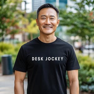 Puede incluir: Una camiseta negra con el texto "DESK JOCKEY" en blanco, usada por una persona sonriente. La persona está afuera con un fondo borroso de árboles y edificios.