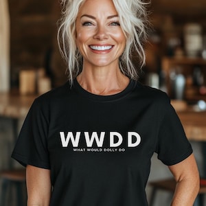 Pode incluir: Camiseta preta com o texto branco "WWDD WHAT WOULD DOLLY DO". A pessoa que usa a camiseta tem cabelo loiro e está sorrindo. O fundo é um interior desfocado.