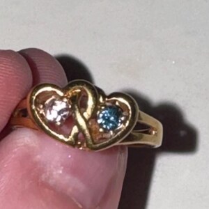 May include: A gold-toned ring featuring two interlocking heart-shaped settings. The left heart contains a pale pink round gemstone, while the right heart holds a deep blue round gemstone. The band is split into two thin sections on each side.