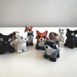 May include: A collection of small, colorful animal figurines. The assortment includes a black cat, white cat, fox, raccoon, and other creatures. Each figure has detailed features and is set against a plain white background.