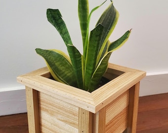 May include: A square wooden planter box with a snake plant. The planter is made of light-colored wood and has a simple, modern design. The snake plant has green and yellow striped leaves, adding a pop of color to the natural wood.