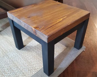 May include: A small, square wooden footstool with a stained brown top and black legs. The footstool is placed on a beige rug with a gray border. The wood grain is visible on the top surface.