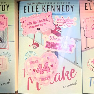 Puede incluir: Una pila de libros de Elle Kennedy. El libro central tiene pegatinas rosas que dicen "Lessons on Ice, Problems Off It", "Briar Hockey" y "44 Hockey Make". Las portadas de los libros son de color azul claro, rosa y blanco.