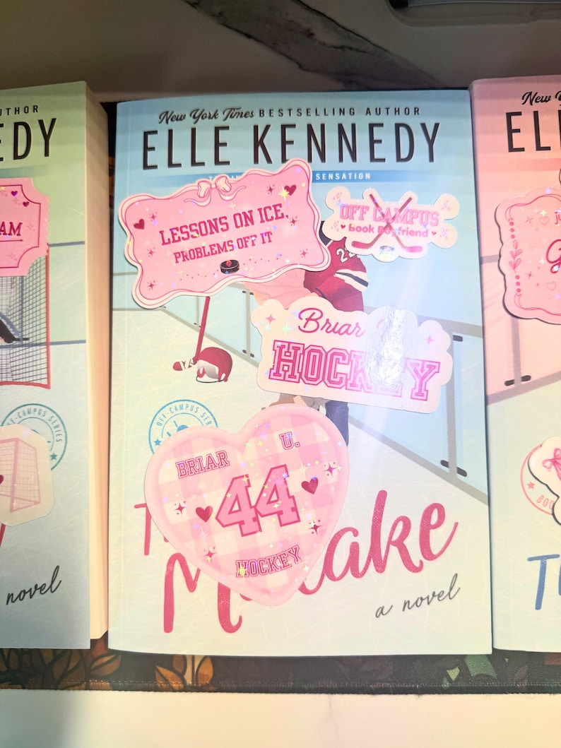 Puede incluir: Una pila de libros de Elle Kennedy. El libro central tiene pegatinas rosas que dicen "Lessons on Ice, Problems Off It", "Briar Hockey" y "44 Hockey Make". Las portadas de los libros son de color azul claro, rosa y blanco.