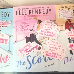 Puede incluir: Imagen de tres libros de Elle Kennedy, incluyendo "The Score". Pegatinas rosas y blancas con frases como "Puck Bunny" y "I like my hockey players" est&aacute;n colocadas en los libros. En el fondo, se ve un estuche transparente con pinzas.