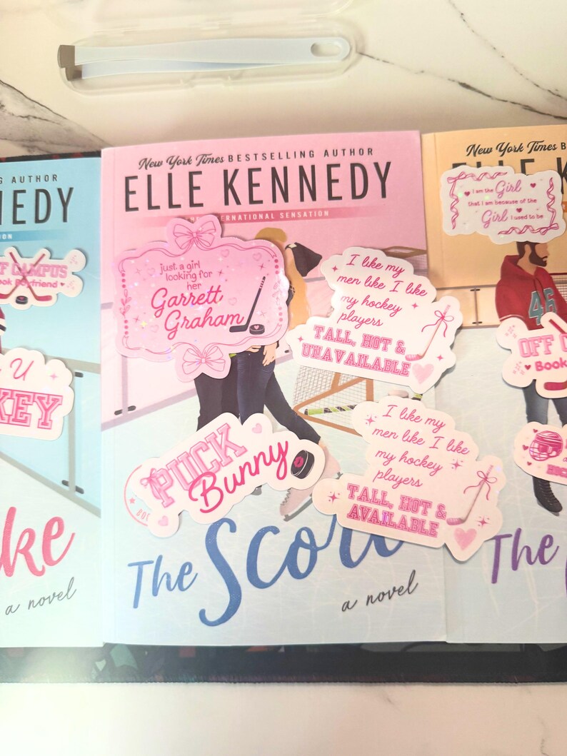 Puede incluir: Imagen de tres libros de Elle Kennedy, incluyendo "The Score". Pegatinas rosas y blancas con frases como "Puck Bunny" y "I like my hockey players" est&aacute;n colocadas en los libros. En el fondo, se ve un estuche transparente con pinzas.