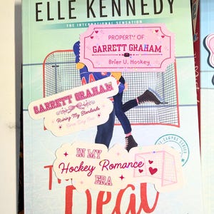 Puede incluir: Portada del libro "The Deal" de Elle Kennedy, con tem&aacute;tica de hockey. La portada tiene un fondo azul claro con una ilustraci&oacute;n de jugador de hockey y el t&iacute;tulo en rojo. Pegatinas dicen "Property of Garrett Graham".