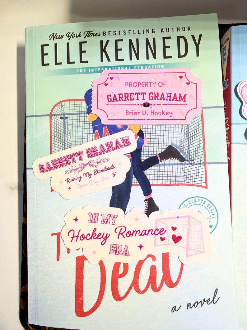 Puede incluir: Portada del libro "The Deal" de Elle Kennedy, con tem&aacute;tica de hockey. La portada tiene un fondo azul claro con una ilustraci&oacute;n de jugador de hockey y el t&iacute;tulo en rojo. Pegatinas dicen "Property of Garrett Graham".