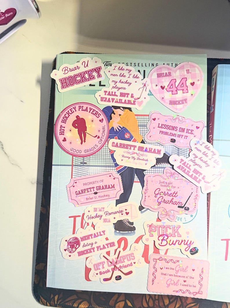 Puede incluir: Una colecci&oacute;n de pegatinas rosas y blancas con temas de hockey, que incluyen corazones, palos de hockey y texto como "Hockey Players" y "Tall, Hot & Unavailable". Las pegatinas est&aacute;n dispuestas sobre un libro.