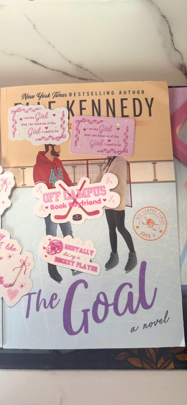 Puede incluir: Una novela titulada "The Goal" de Elle Kennedy, con pegatinas decorativas. Las pegatinas presentan frases como "Off Campus Book Boyfriend" y "Mentally doing a hockey player". La portada del libro tiene un dise&ntilde;o de pista de hockey.