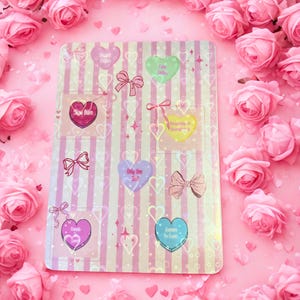 May include: A pink and white striped notepad with heart-shaped stickers and bows. The stickers have text such as "Sweet Daze", "Slow Burn", and "Friends". The background is covered in pink roses.