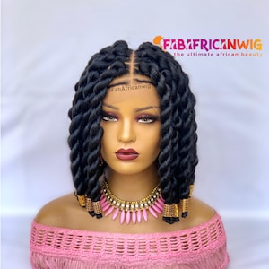 May include: A black braided wig with a center part, styled on a mannequin head. The wig features thick, twisted braids and is adorned with gold accents at the ends. The mannequin wears a pink off-the-shoulder top and a pink and gold necklace. The logo "FabAfricanWig" is visible.