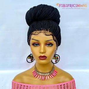 May include: A black braided wig styled in a high bun, with a lace front. The model is wearing a pink off-the-shoulder top, a pink and gold necklace, and black earrings with an African continent design. The text "FAB AFRICAN WIG" is visible.