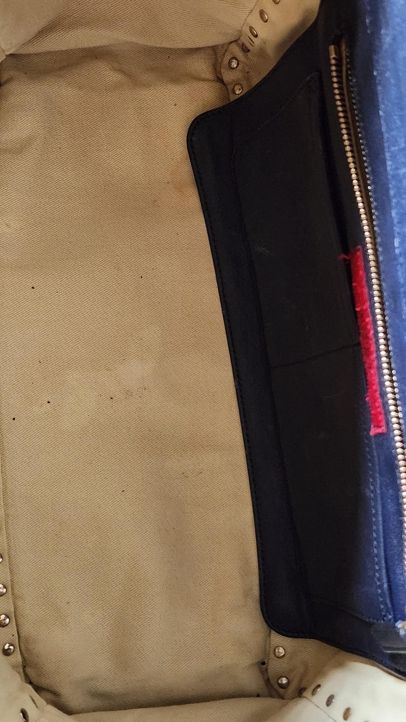 May include: Interior view of a handbag, featuring a beige fabric lining and a navy blue leather pocket with a red accent. The bag's interior shows signs of wear.