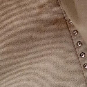 May include: Close-up of a beige fabric with a diagonal weave. The fabric has some darker spots and a row of silver-colored buttons along the edge. The texture of the material is visible.