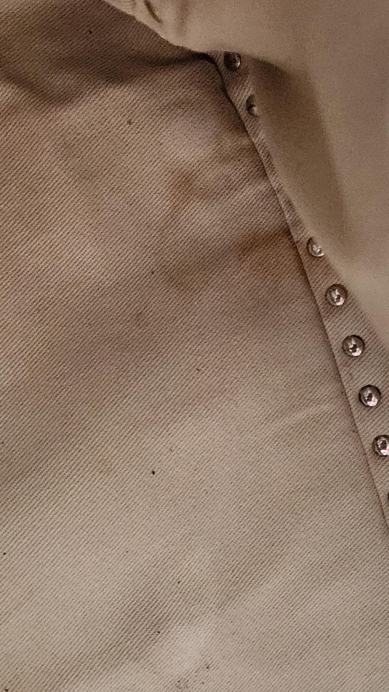 May include: Close-up of a beige fabric with a diagonal weave. The fabric has some darker spots and a row of silver-colored buttons along the edge. The texture of the material is visible.