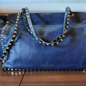 May include: A navy blue leather handbag with gold-colored pyramid studs along the edges and straps. The bag has a shoulder strap and a top handle. The Valentino logo is visible on the front of the bag.