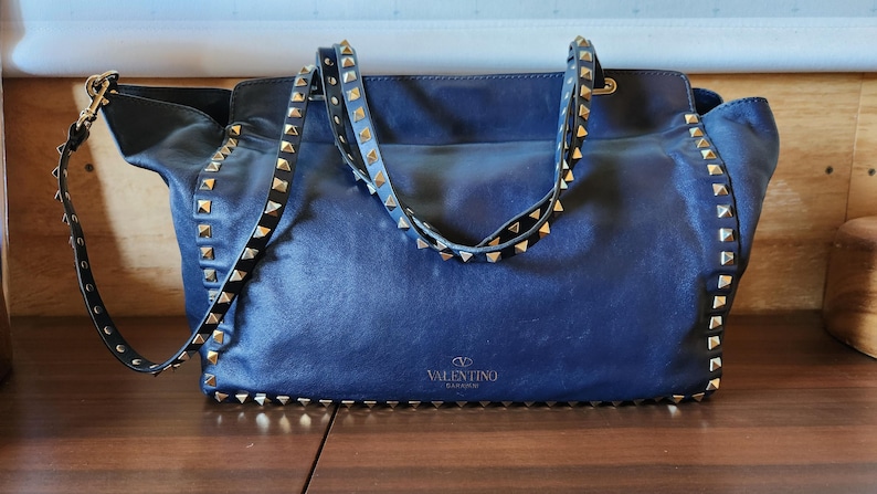 May include: A navy blue leather handbag with gold-colored pyramid studs along the edges and straps. The bag has a shoulder strap and a top handle. The Valentino logo is visible on the front of the bag.