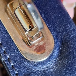 May include: Close-up of a gold-toned metal clasp on a navy blue leather surface. The clasp is rectangular with a curved top and a silver-colored lever. The leather has visible stitching and a textured appearance.