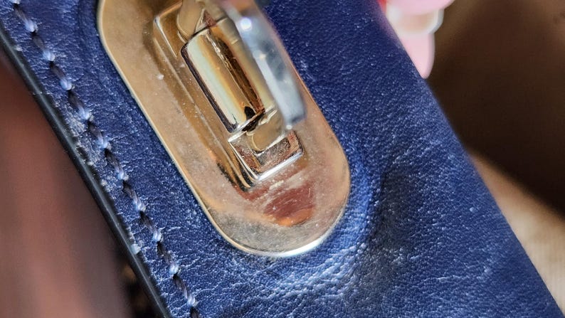 May include: Close-up of a gold-toned metal clasp on a navy blue leather surface. The clasp is rectangular with a curved top and a silver-colored lever. The leather has visible stitching and a textured appearance.