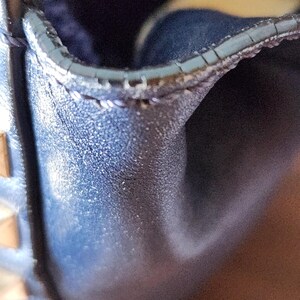 May include: Close-up of a section of a dark blue leather bag. The image highlights the textured surface and stitching along the edge. Gold-colored studs are visible on the side. The bag appears to be well-crafted and stylish.