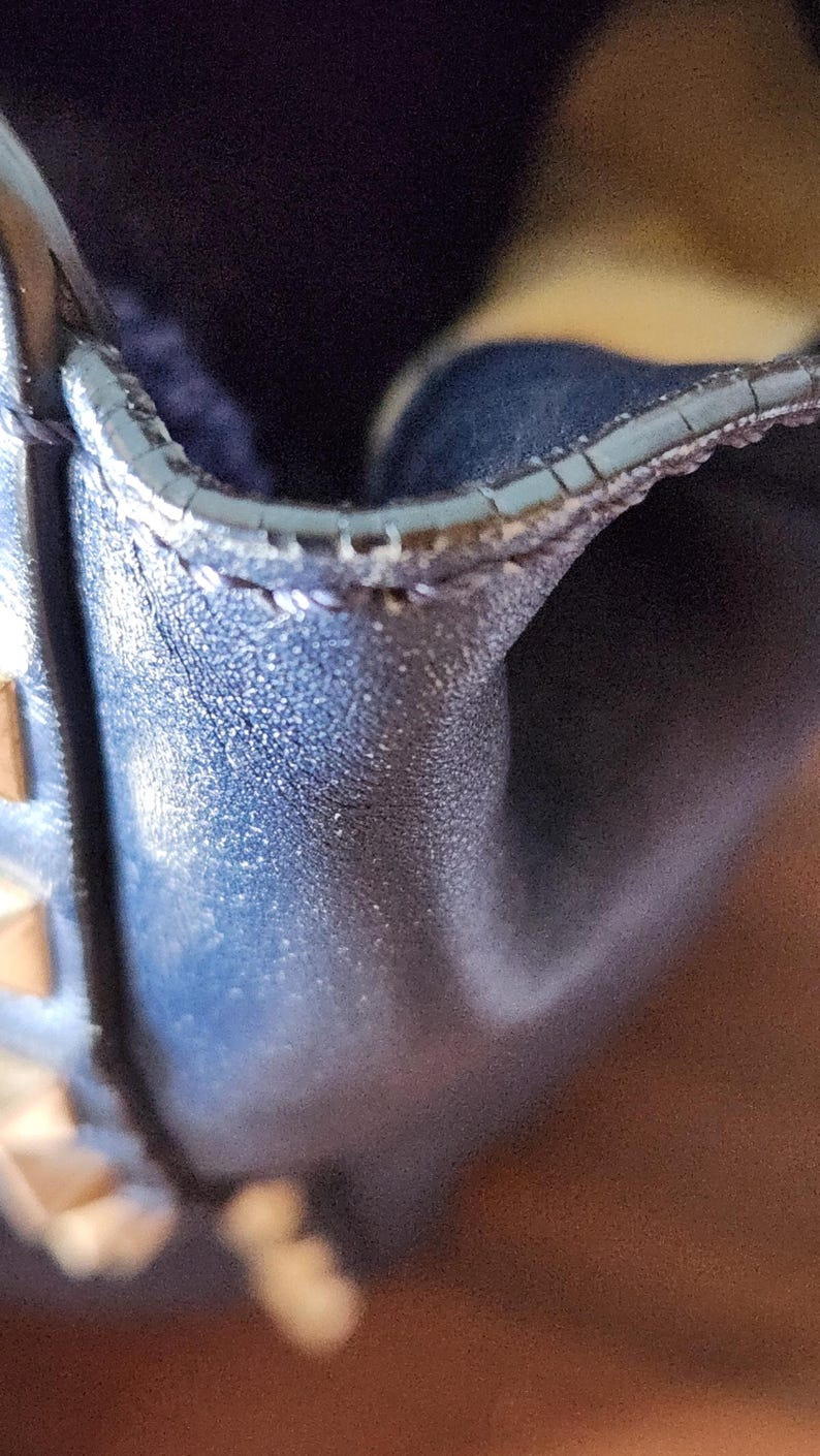 May include: Close-up of a section of a dark blue leather bag. The image highlights the textured surface and stitching along the edge. Gold-colored studs are visible on the side. The bag appears to be well-crafted and stylish.