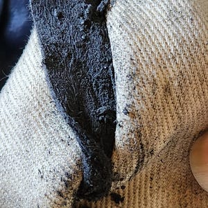 May include: Close-up of a torn fabric with a dark, frayed edge. The fabric appears to be a light beige color, with a visible weave. The tear reveals a dark, possibly black, inner layer.