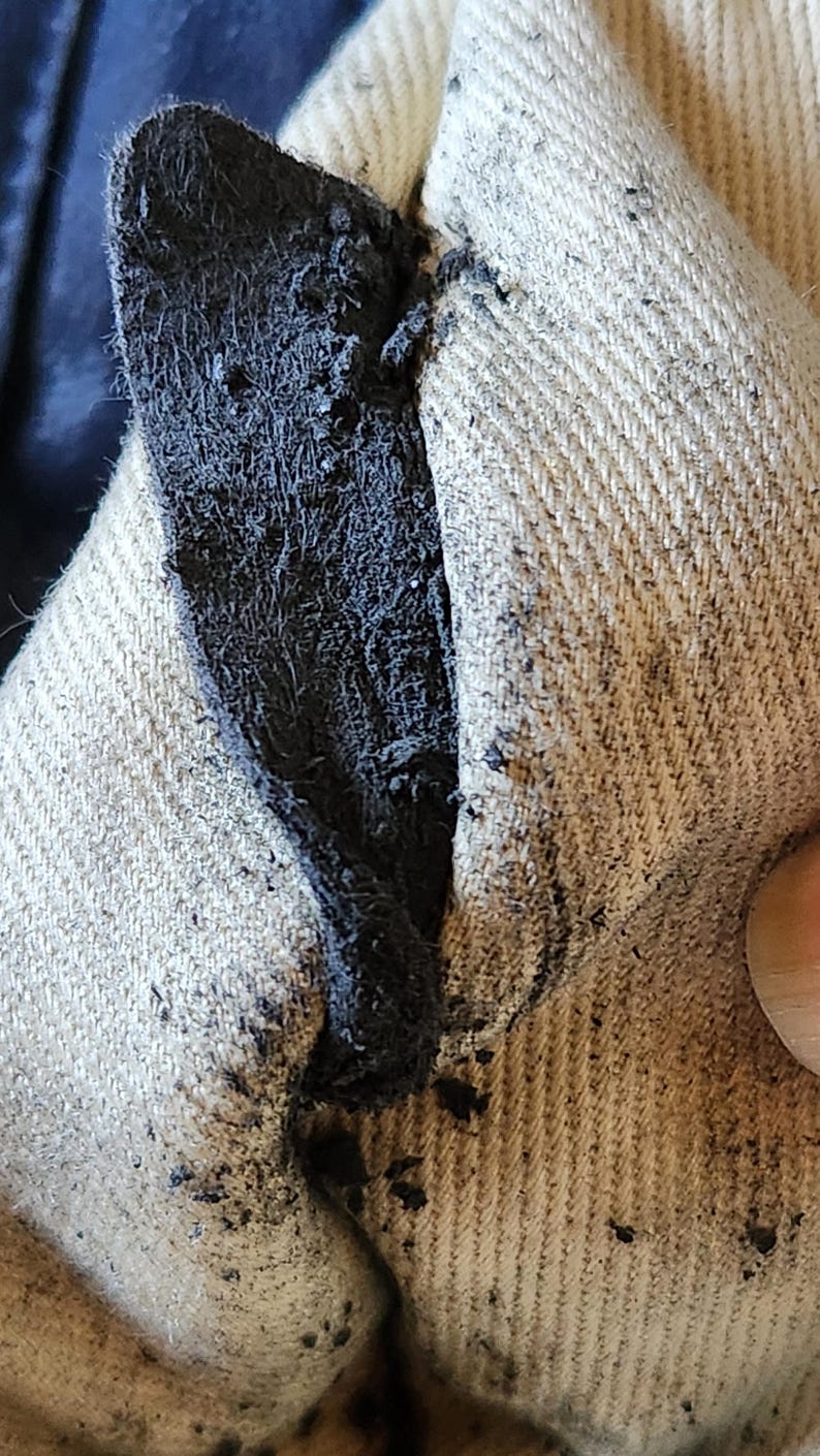 May include: Close-up of a torn fabric with a dark, frayed edge. The fabric appears to be a light beige color, with a visible weave. The tear reveals a dark, possibly black, inner layer.