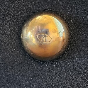 May include: Close-up of a gold-colored, rounded stud with a small, engraved design, set against a textured black leather surface. The stud reflects light, creating highlights. The leather has a subtle grain.