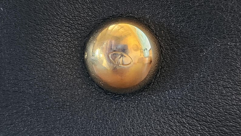 May include: Close-up of a gold-colored, rounded stud with a small, engraved design, set against a textured black leather surface. The stud reflects light, creating highlights. The leather has a subtle grain.