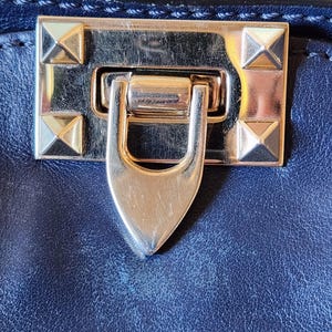 May include: Close-up of a navy blue leather handbag clasp. The gold-toned metal clasp features a rectangular plate with four pyramid studs and a curved, pointed latch. The bag's leather has a textured appearance.