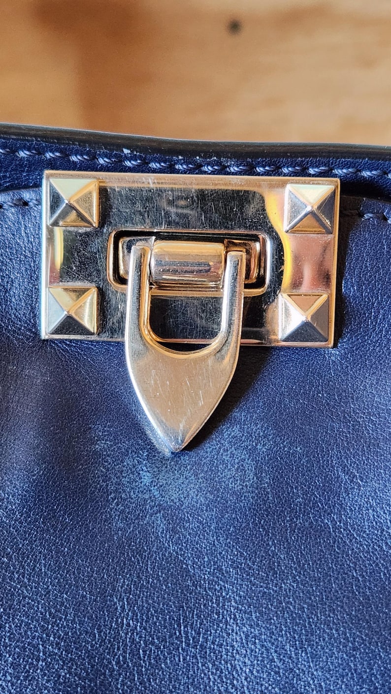 May include: Close-up of a navy blue leather handbag clasp. The gold-toned metal clasp features a rectangular plate with four pyramid studs and a curved, pointed latch. The bag's leather has a textured appearance.