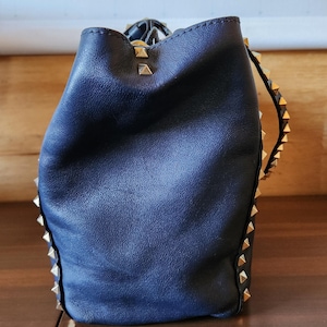 May include: A navy blue leather bucket bag with gold-colored pyramid studs along the edges. The bag has a drawstring closure and a soft, slouchy shape. The background includes a map of Antarctica.
