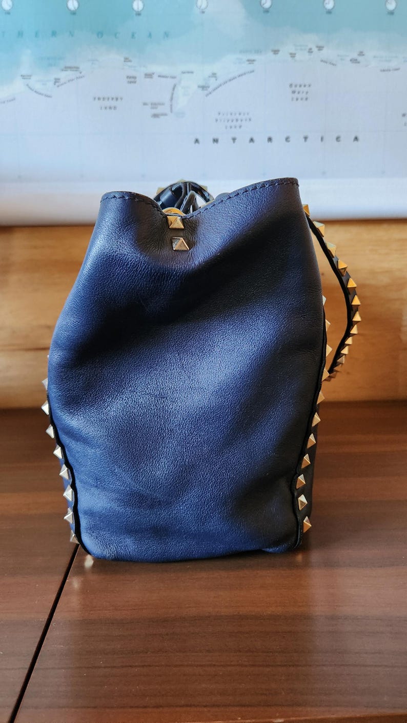 May include: A navy blue leather bucket bag with gold-colored pyramid studs along the edges. The bag has a drawstring closure and a soft, slouchy shape. The background includes a map of Antarctica.