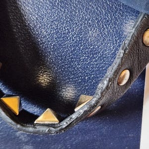 May include: Close-up of a navy blue leather handbag with a black strap. The strap is adorned with gold pyramid studs and gold round studs. The bag has a textured leather surface.