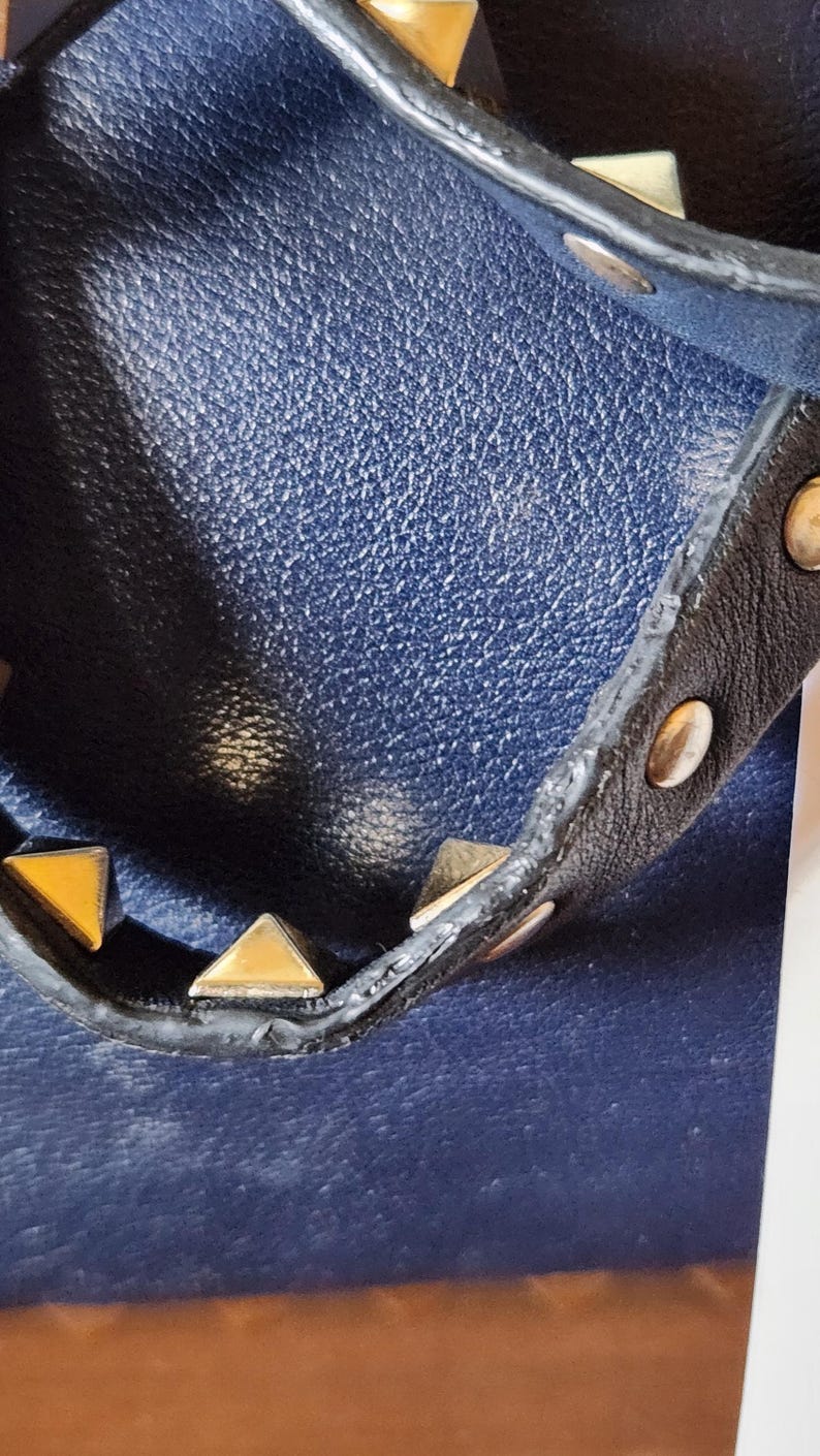 May include: Close-up of a navy blue leather handbag with a black strap. The strap is adorned with gold pyramid studs and gold round studs. The bag has a textured leather surface.