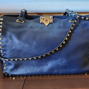 May include: A navy blue leather handbag with gold-colored pyramid studs along the edges and straps. The bag has a top handle and a shoulder strap. The clasp is gold-colored.