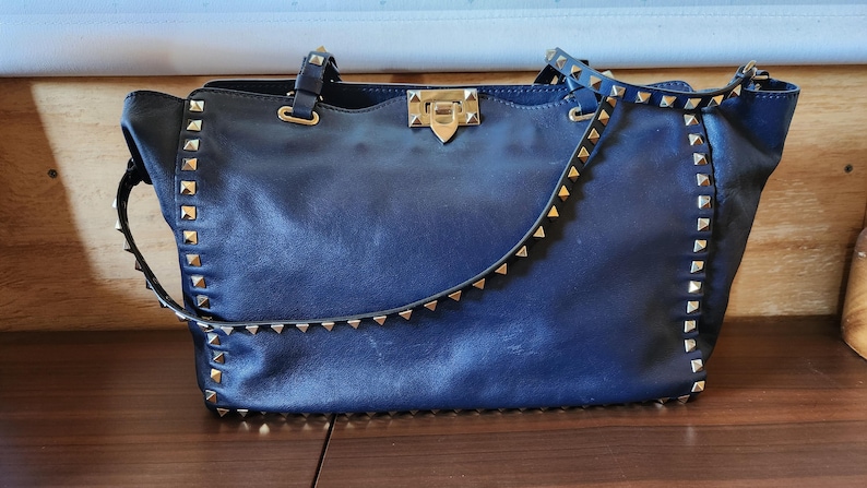 May include: A navy blue leather handbag with gold-colored pyramid studs along the edges and straps. The bag has a top handle and a shoulder strap. The clasp is gold-colored.