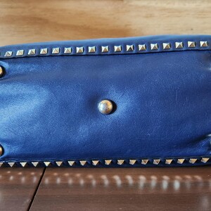 May include: A navy blue leather handbag with silver-colored pyramid studs along the edges and four round silver-colored feet. The bottom of the bag is visible, resting on a wooden surface.
