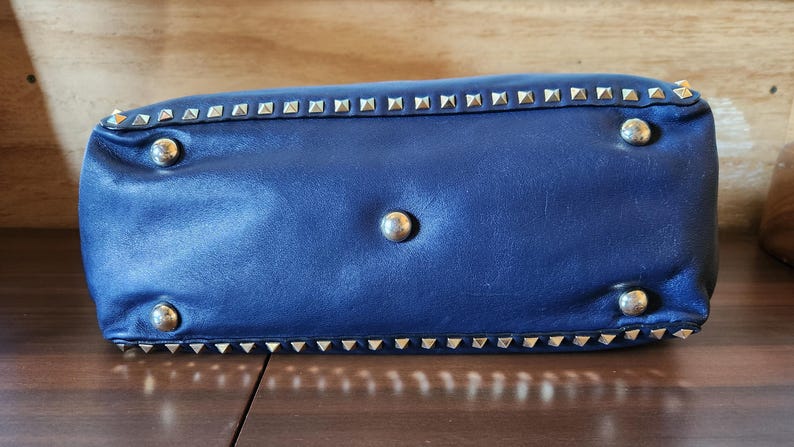May include: A navy blue leather handbag with silver-colored pyramid studs along the edges and four round silver-colored feet. The bottom of the bag is visible, resting on a wooden surface.