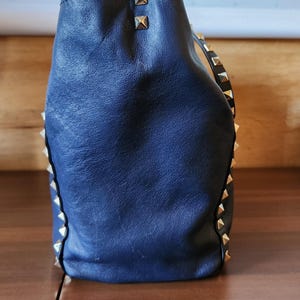 May include: A navy blue leather handbag with gold-colored pyramid studs along the edges and top. The bag has a soft, slouchy shape and a textured surface. The bag is sitting on a wooden surface.