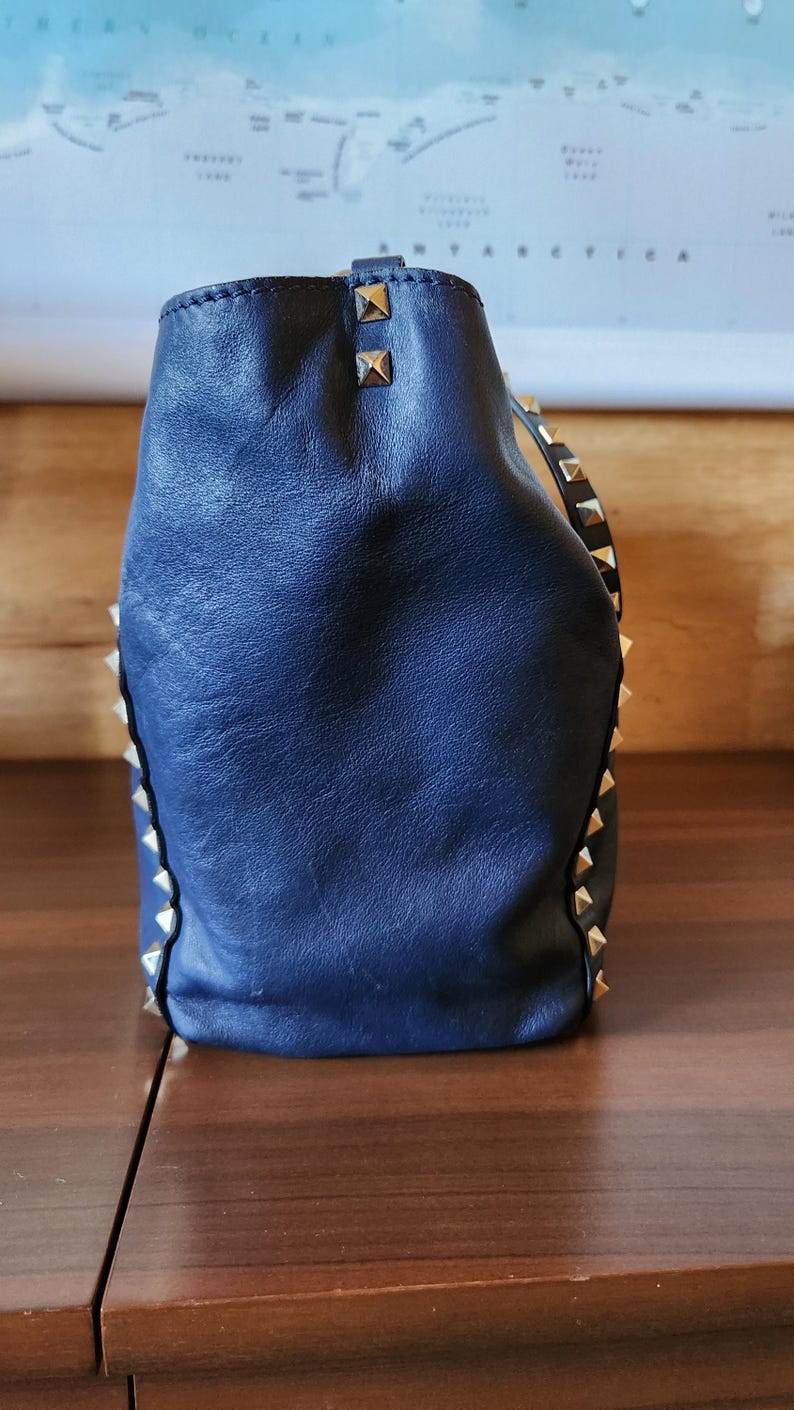 May include: A navy blue leather handbag with gold-colored pyramid studs along the edges and top. The bag has a soft, slouchy shape and a textured surface. The bag is sitting on a wooden surface.