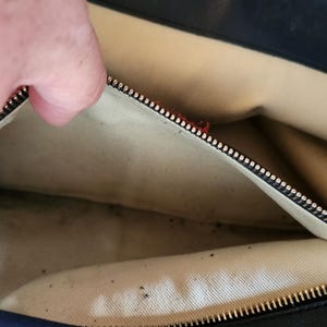 May include: Open view of a black leather handbag interior. The beige lining is visible, with a zipper closure and a hand partially in view. The bag appears to be well-worn.