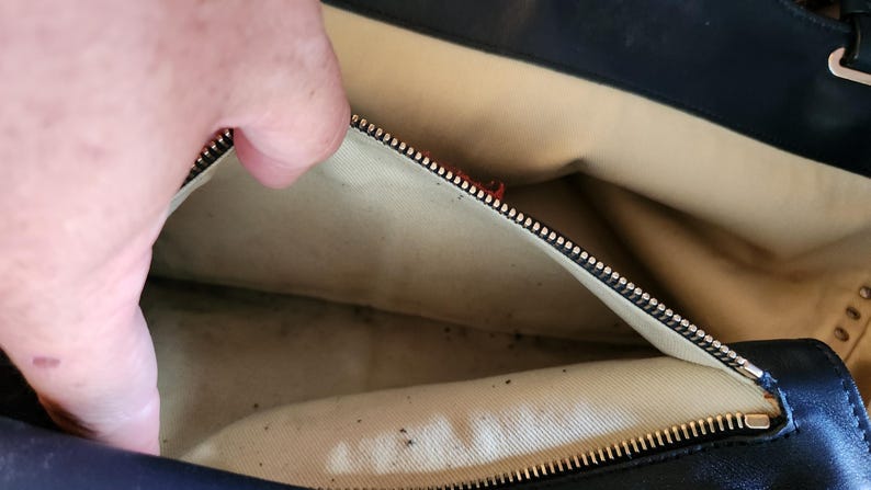 May include: Open view of a black leather handbag interior. The beige lining is visible, with a zipper closure and a hand partially in view. The bag appears to be well-worn.