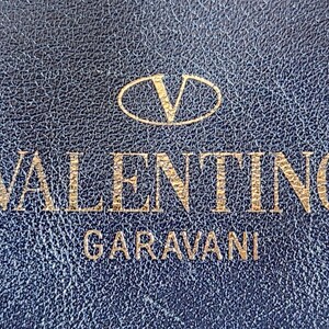 May include: Close-up of a textured, navy blue surface with the gold-colored text "VALENTINO GARAVANI" and a logo. The logo is a gold-colored "V" inside an oval.