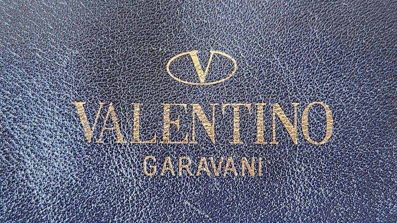 May include: Close-up of a textured, navy blue surface with the gold-colored text "VALENTINO GARAVANI" and a logo. The logo is a gold-colored "V" inside an oval.